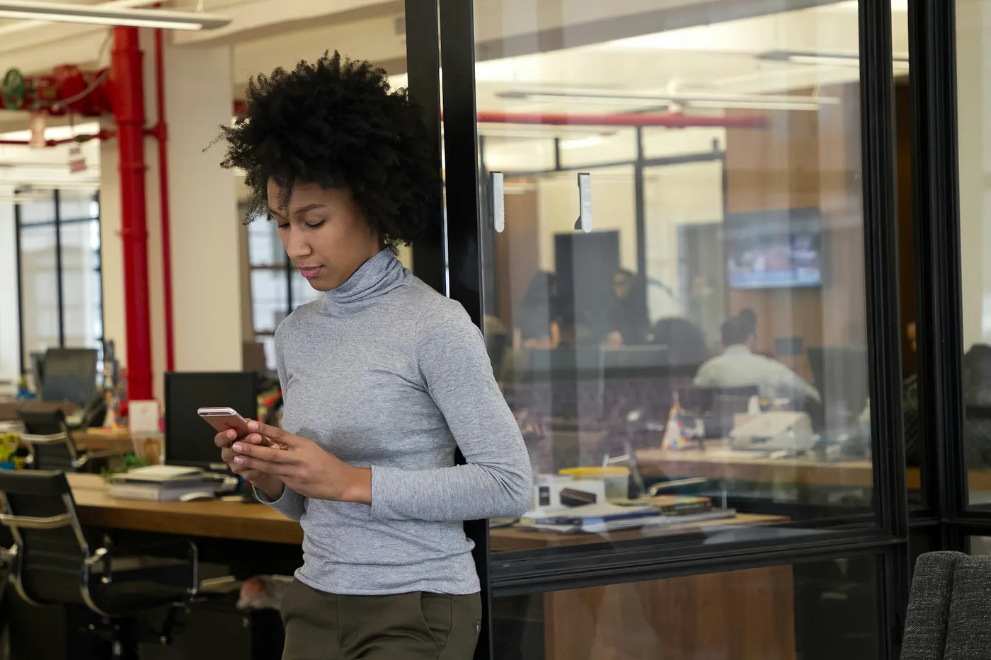 A woman in a grey turtleneck leans against a glass office wall while reading a message on her phone, with colleagues working in a meeting room behind her.