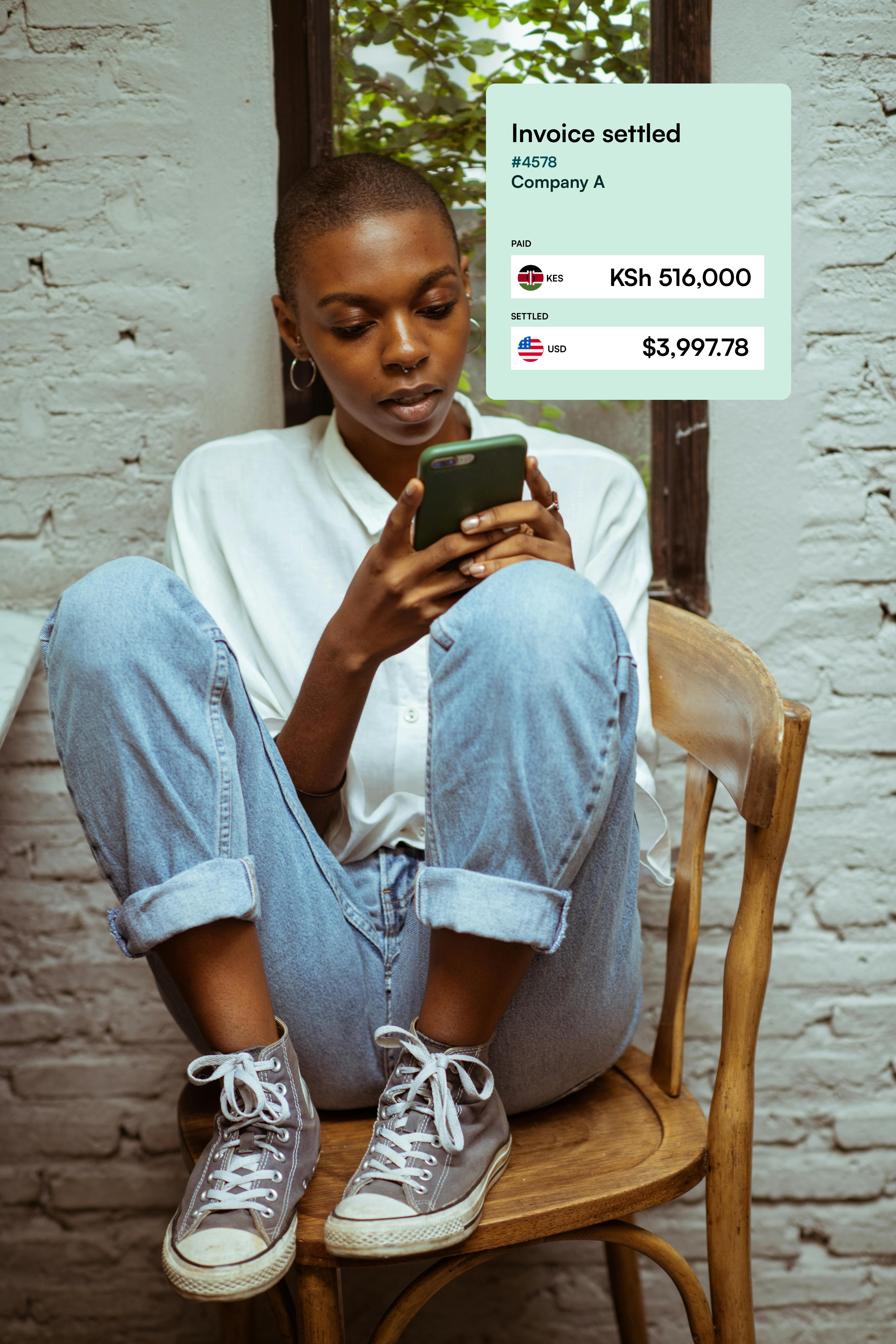 Woman with short hair in white shirt and rolled-up jeans sitting on a wooden chair, looking at her phone next to a window with an invoice graphic showing settled payment of KSh 516,000 and USD 3,997.78.