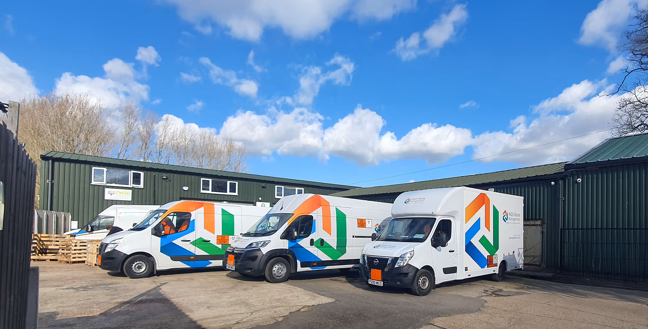 Three brightly colored delivery vans parked in a lot outside a green warehouse building.