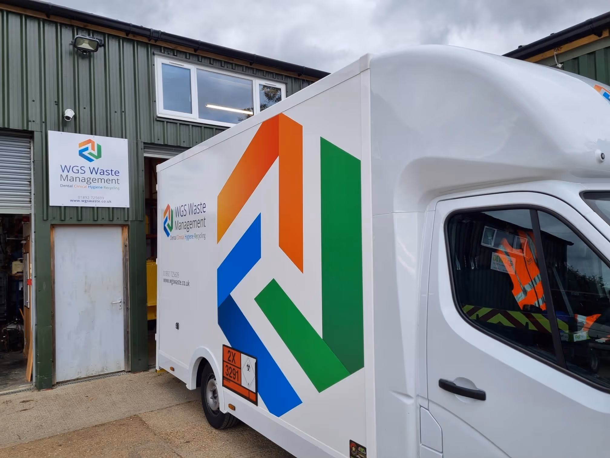 A white waste management truck is parked outside a building with a WGS Waste Management sign.