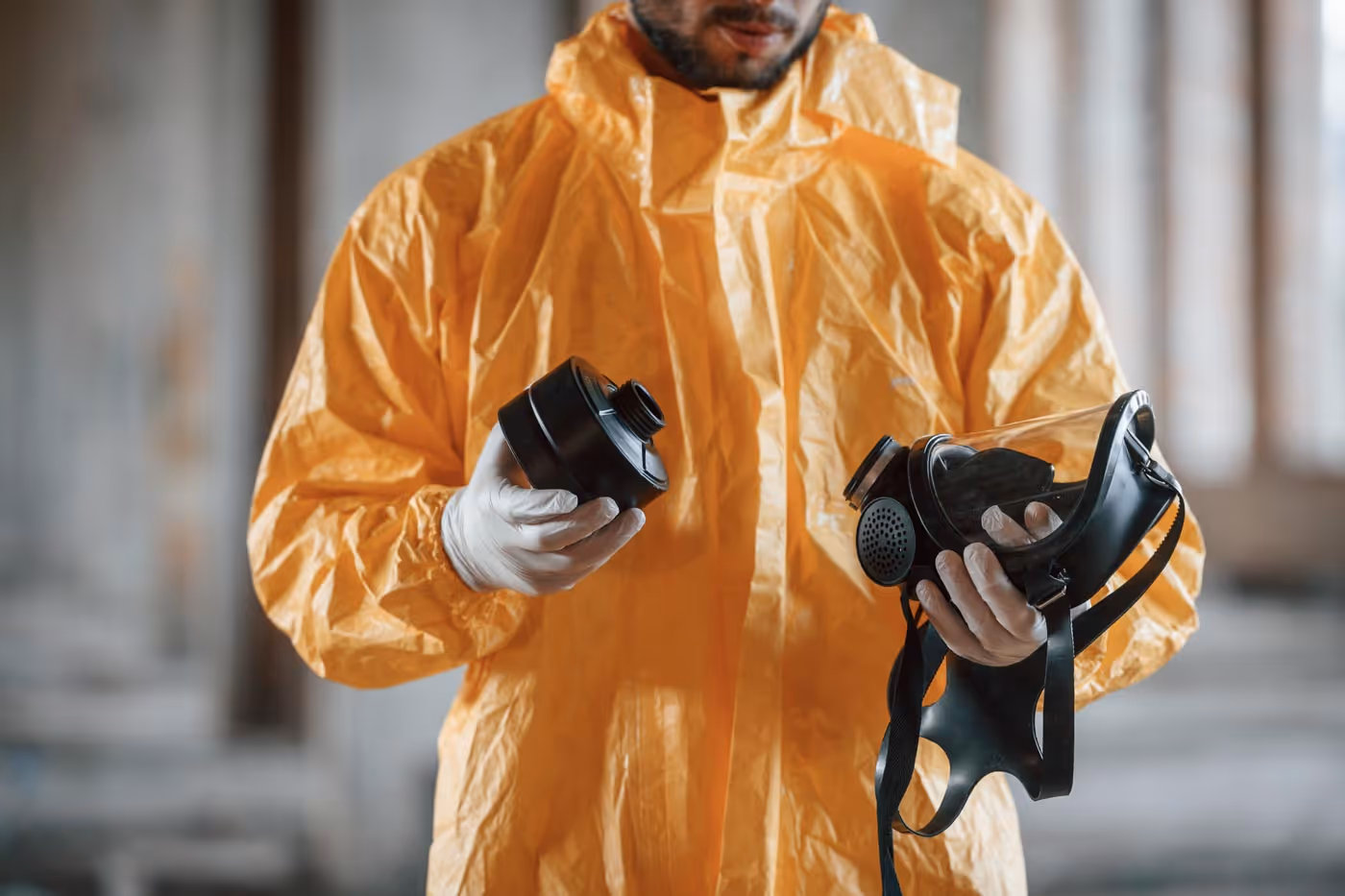 Person in a yellow hazmat suit holding a gas mask and filter inside a building.