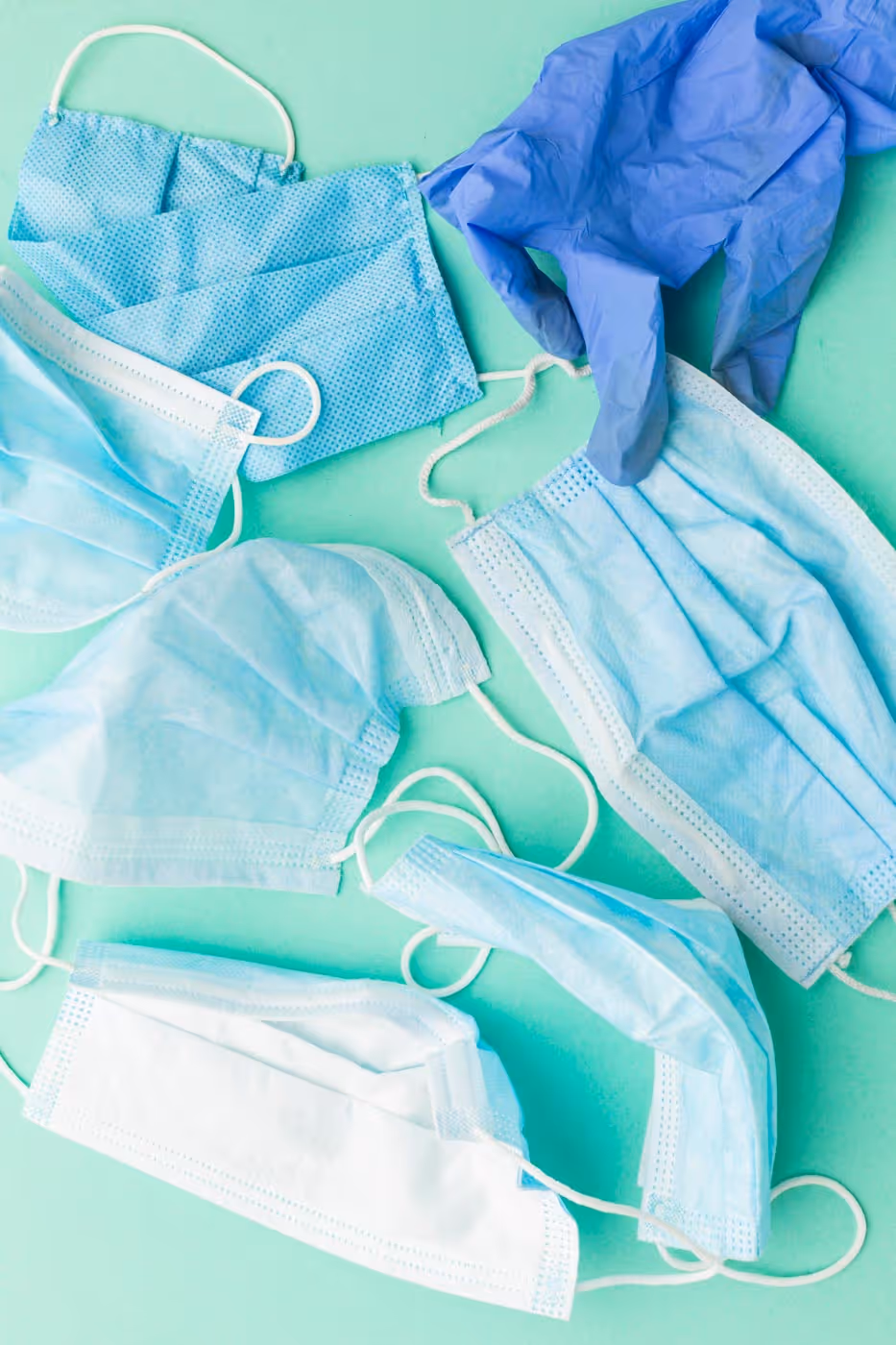 Several blue surgical masks and one blue glove scattered on a light green surface.