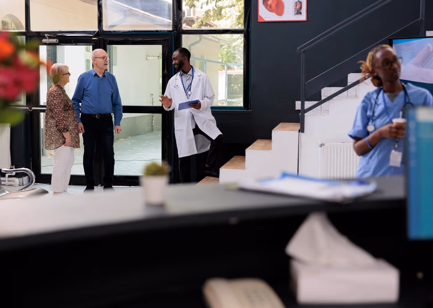 A doctor in a white coat talks to two people in an office hallway.