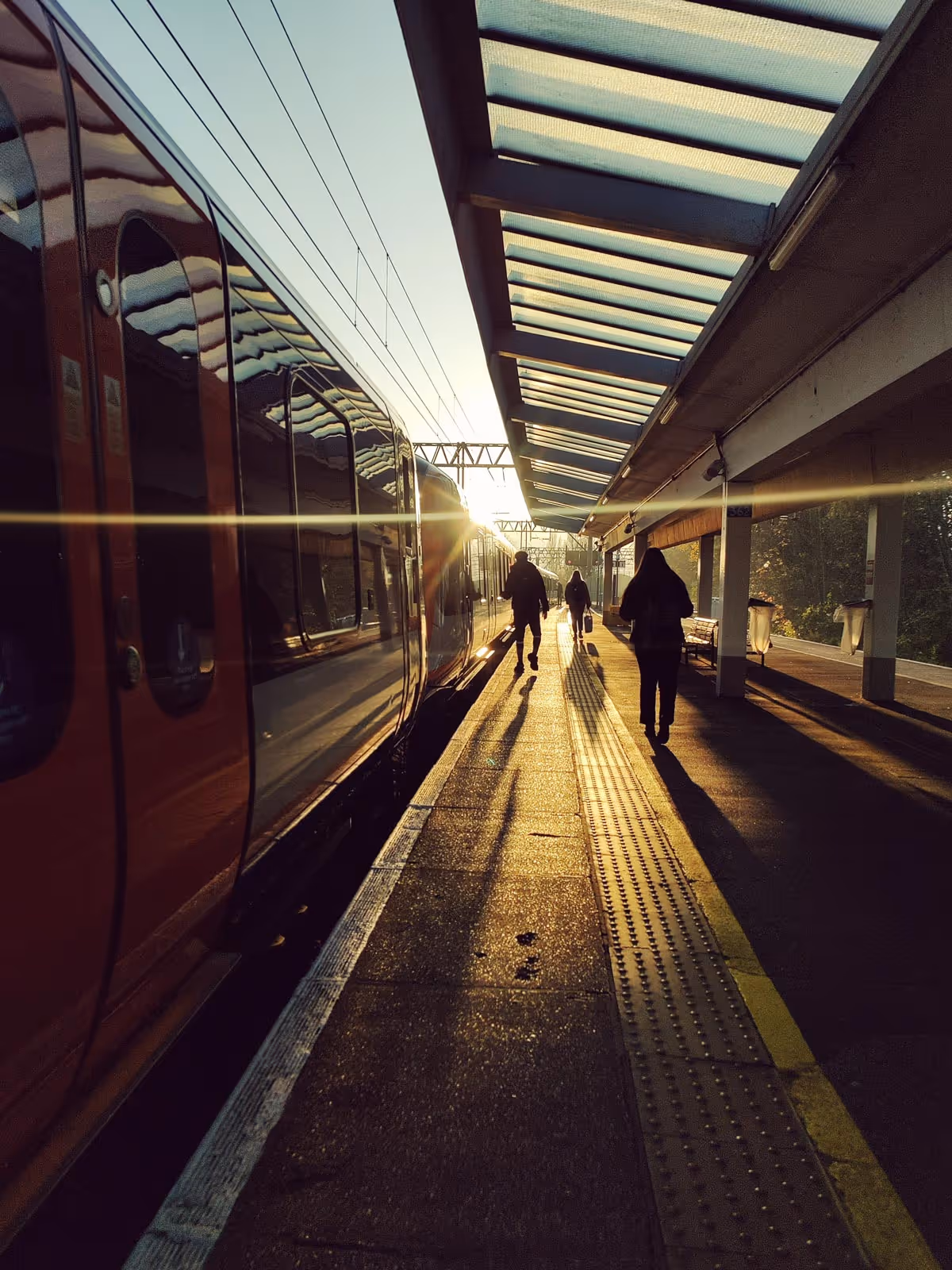 Three people walking along a train platform at sunset, casting long shadows.
