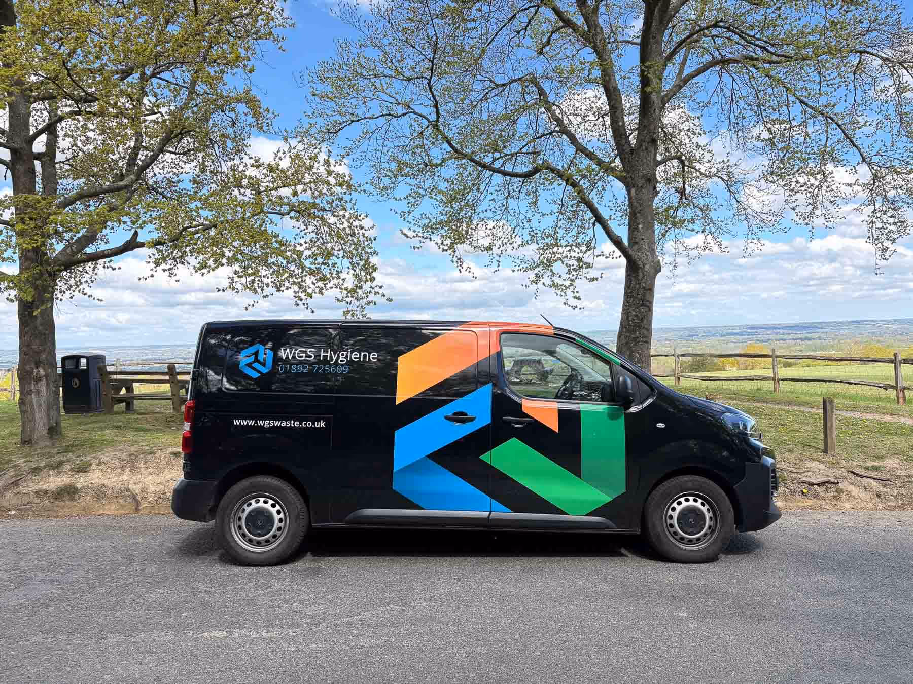 A black van with colorful geometric designs is parked on a street near some trees.