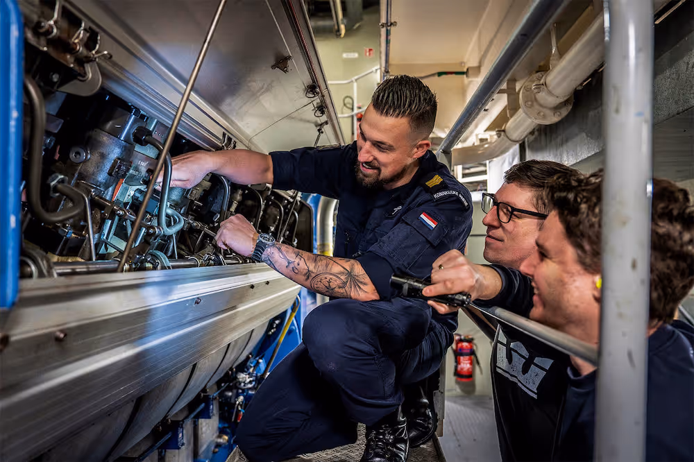 Drie technici werken samen aan een grote machine aan boord, waarbij de middelste met tatoeages en een uniform met Nederlandse vlag een onderdeel aanpast.