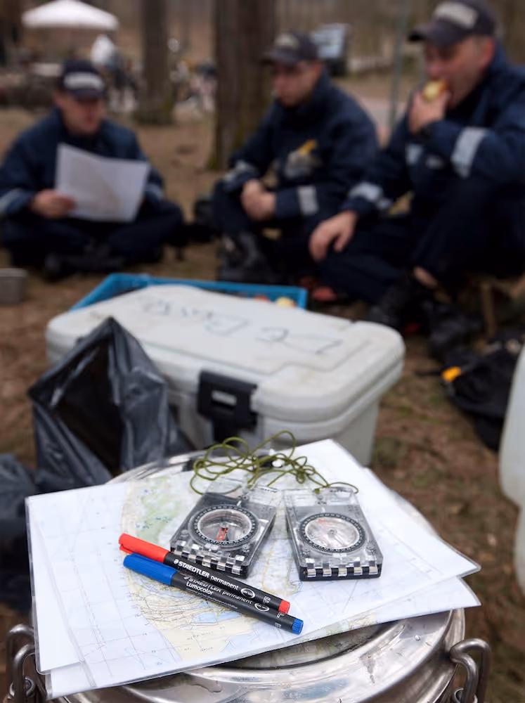 Twee kompasinstrumenten en rode en blauwe stiften op een kaart, met op de achtergrond drie mannen in blauwe uniforme kleding die buiten zitten.