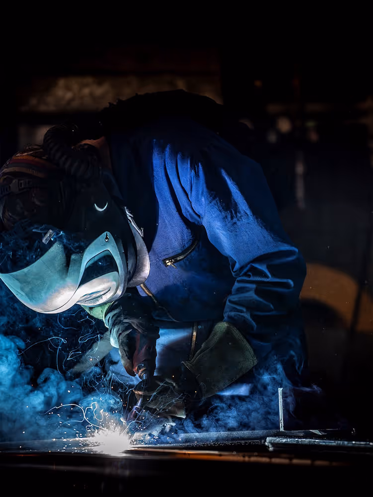 Een lasser in een beschermend pak en helm aan het werk met vonken en rook in een donkere werkplaats.