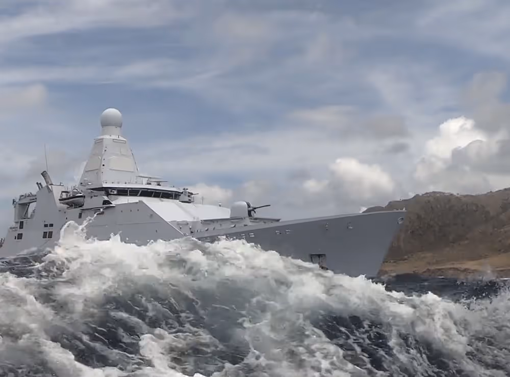 Modern marineschip dat golven breekt op een bewolkte dag dichtbij een rotsachtige kustlijn.