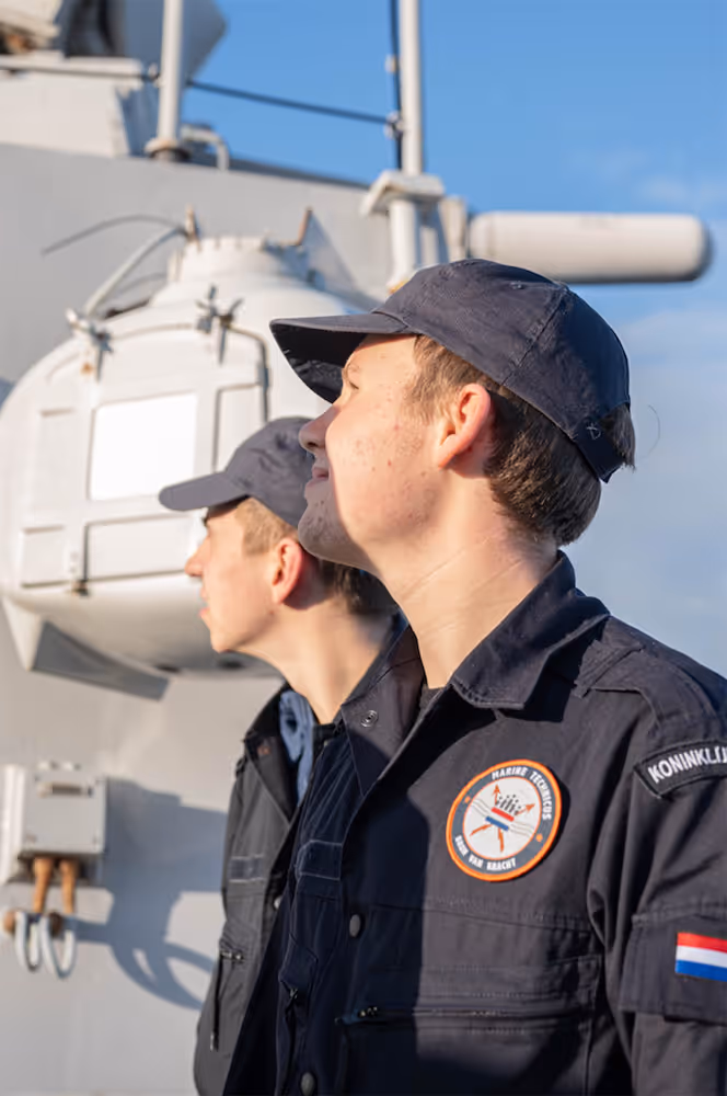 Twee jonge marinetechnici in uniform kijken aandachtig op een marineschip onder een heldere blauwe lucht.