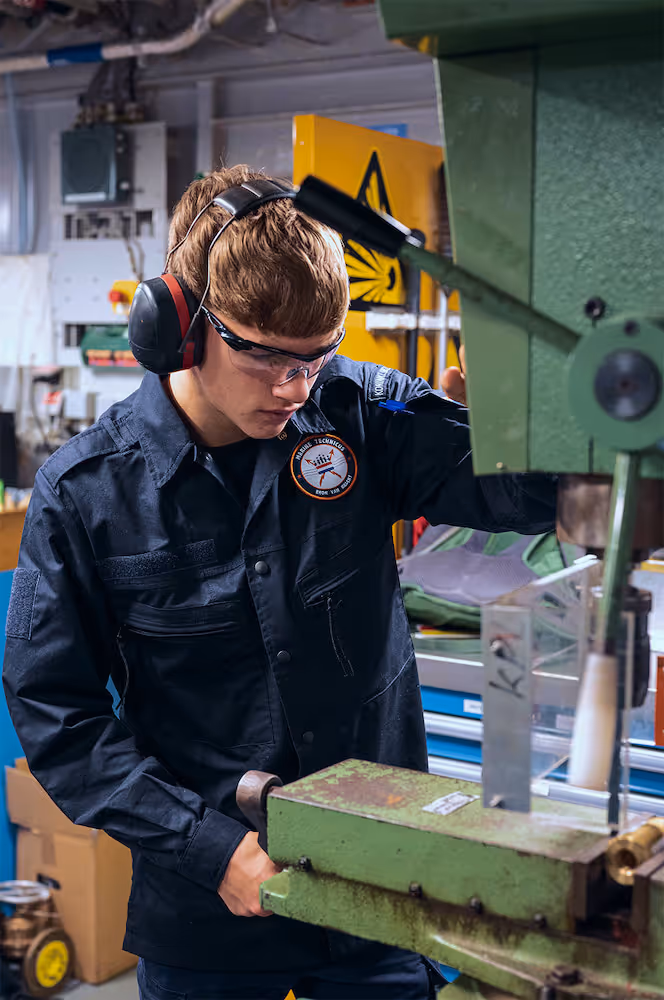 Marine technicus draagt gehoorbescherming en veiligheidsbril terwijl hij een groene industriële machine bedient in een werkplaats.