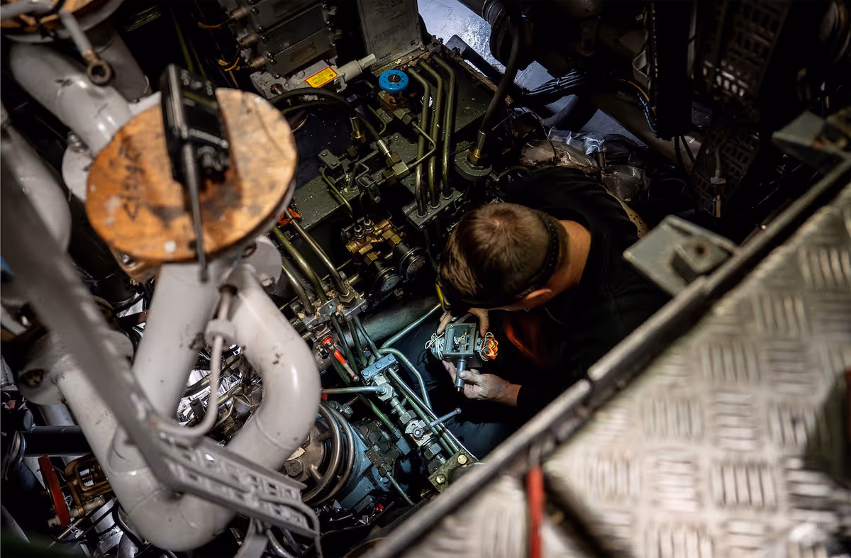 Een monteur werkt aan complexe leidingen en mechanische onderdelen in een machinekamer.