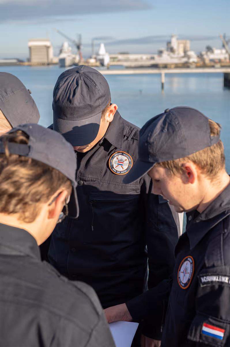 Vier mannen in marine-uniformen met petten overleggen aan dek bij haven met schepen op de achtergrond.