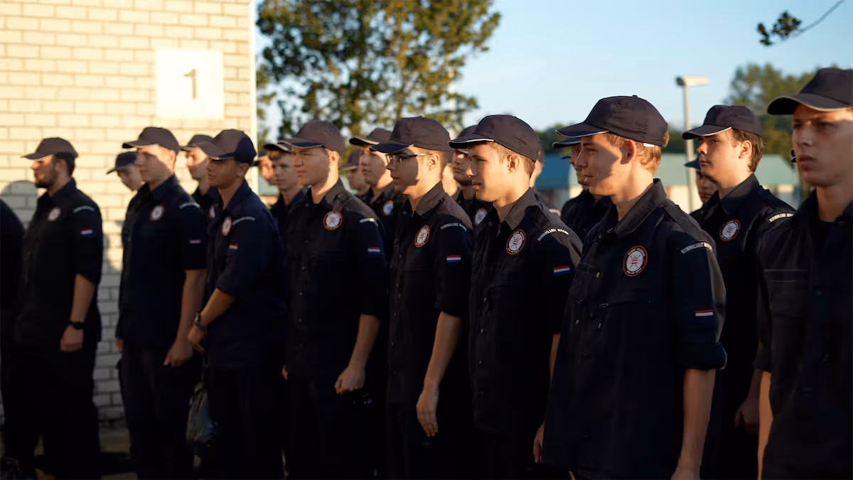 Groep jonge mannen in uniformen en petten die in een rij naast een bakstenen muur staan bij zonlicht.