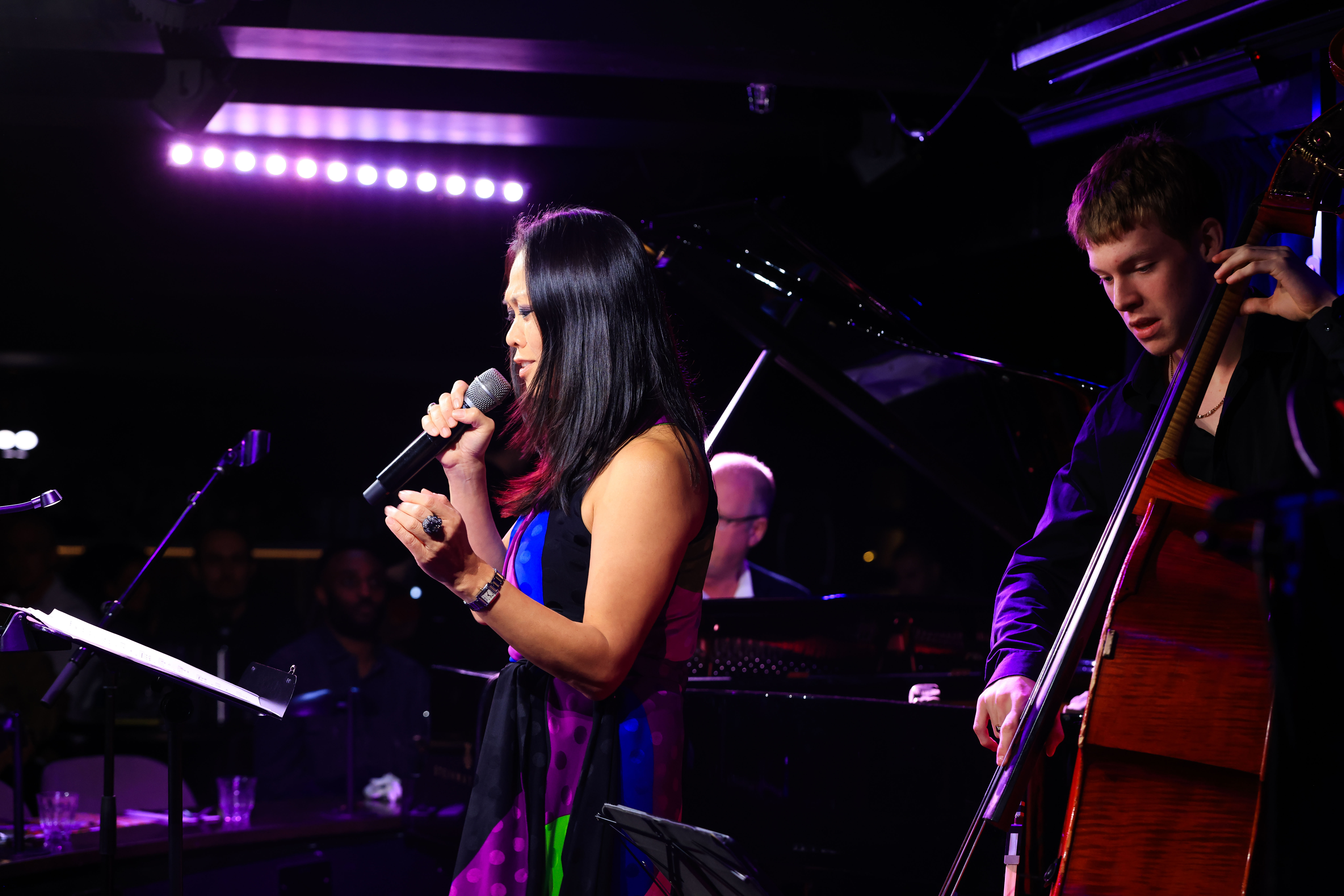 Judy Kawaguchi singer holding a microphone performing on stage beside a male musician playing a double bass, with a pianist in the background.