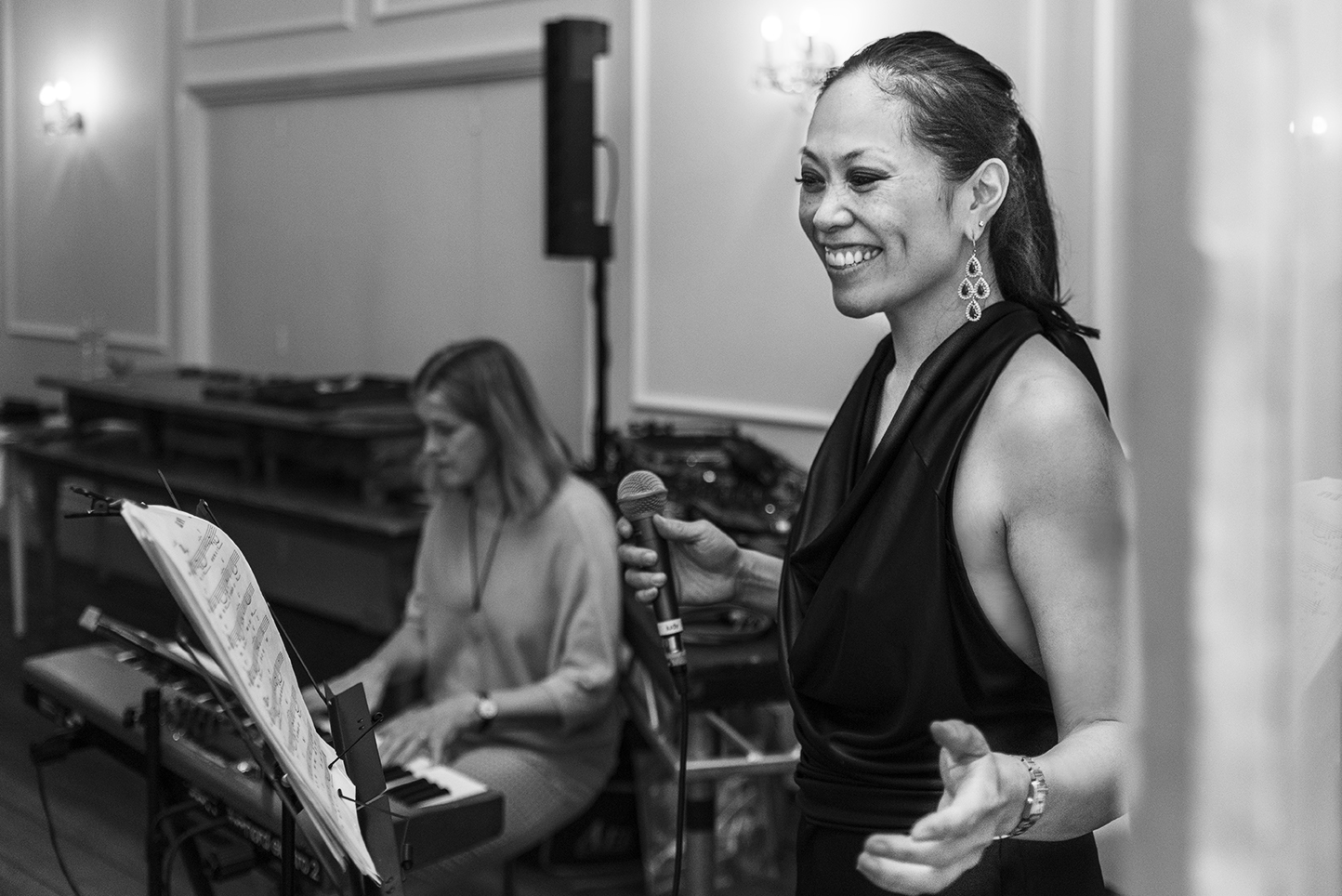 Judy Kawaguchi holding a microphone and gesturing, with a pianist playing keyboard in the background.