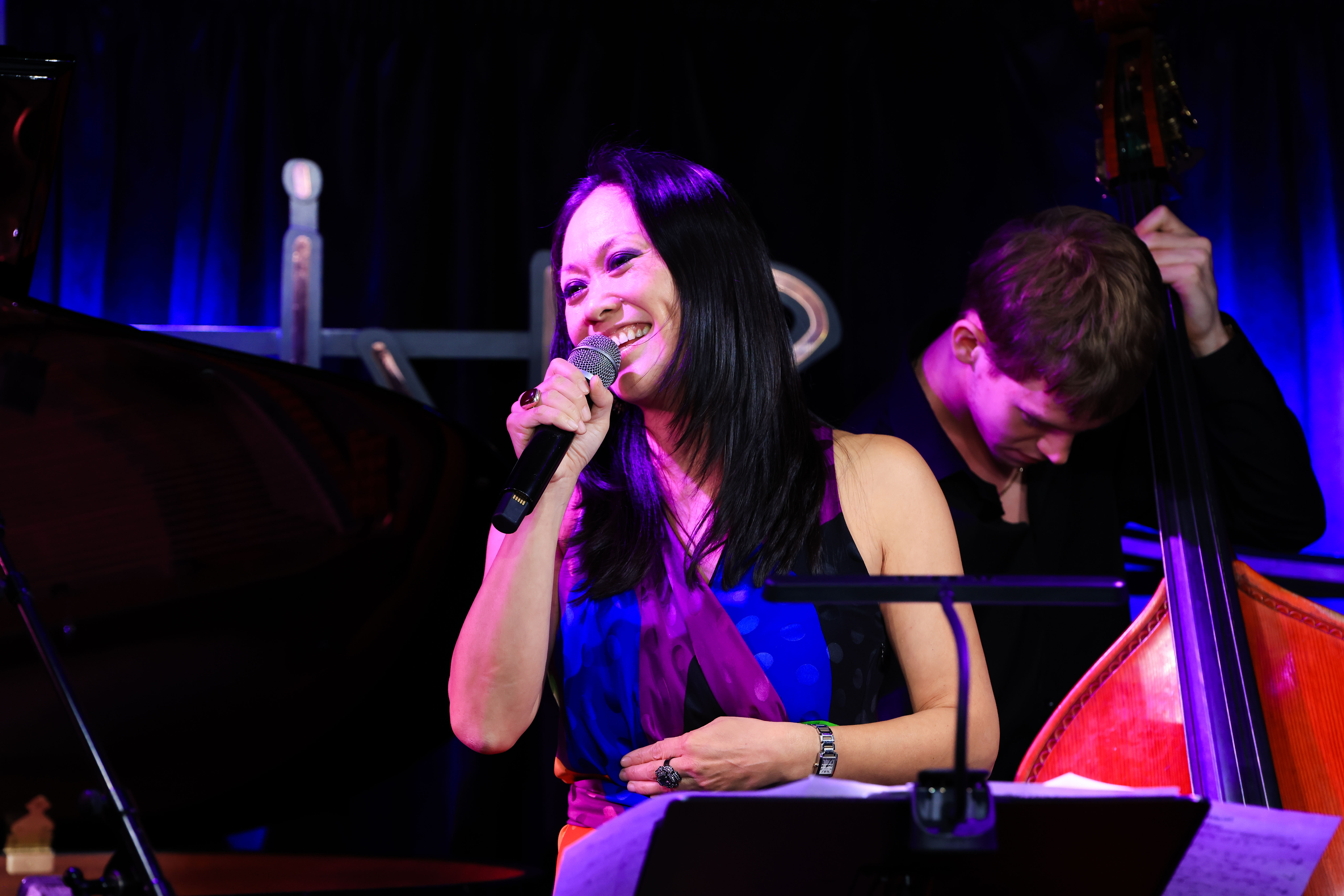 Judy Kawaguchi smiling into microphone with a male double bass player behind her on stage under purple lighting.