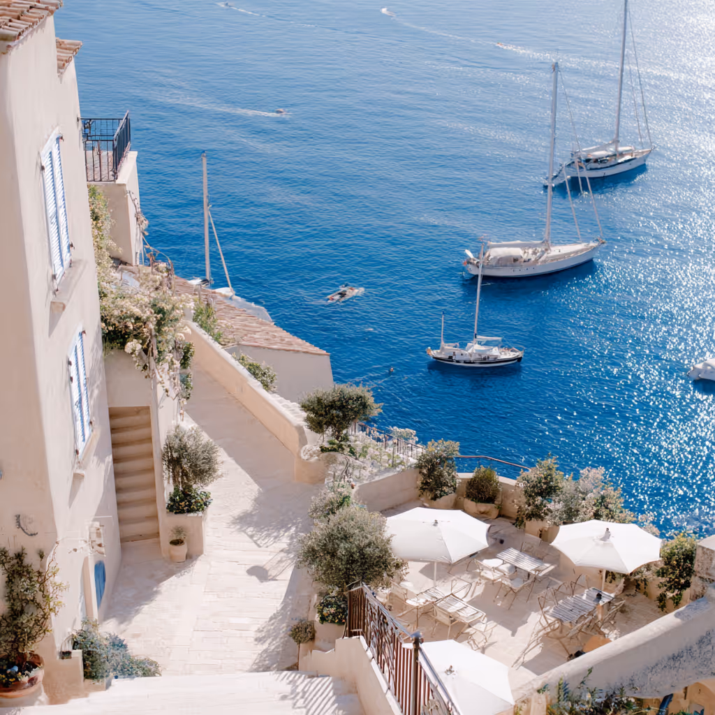 Coastal terrace with white umbrellas and tables overlooking a blue sea with sailboats.