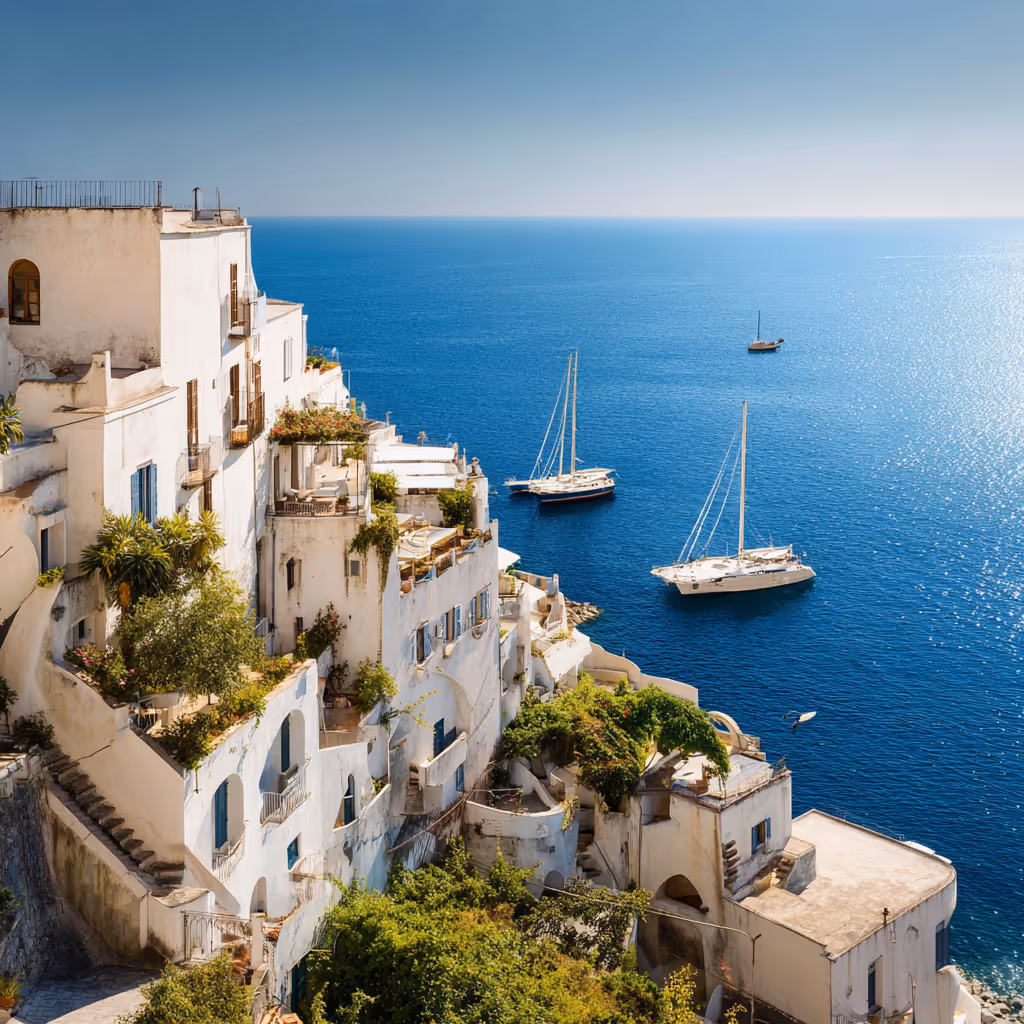 White Mediterranean buildings on a cliffside overlooking a bright blue sea with three sailboats floating nearby.