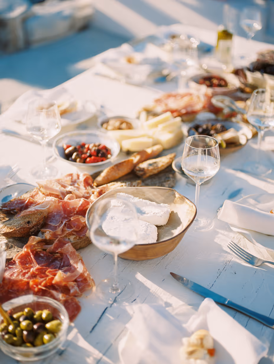 Table set with charcuterie including sliced cured meats, various cheeses, olives, bread, and empty wine glasses in natural daylight.