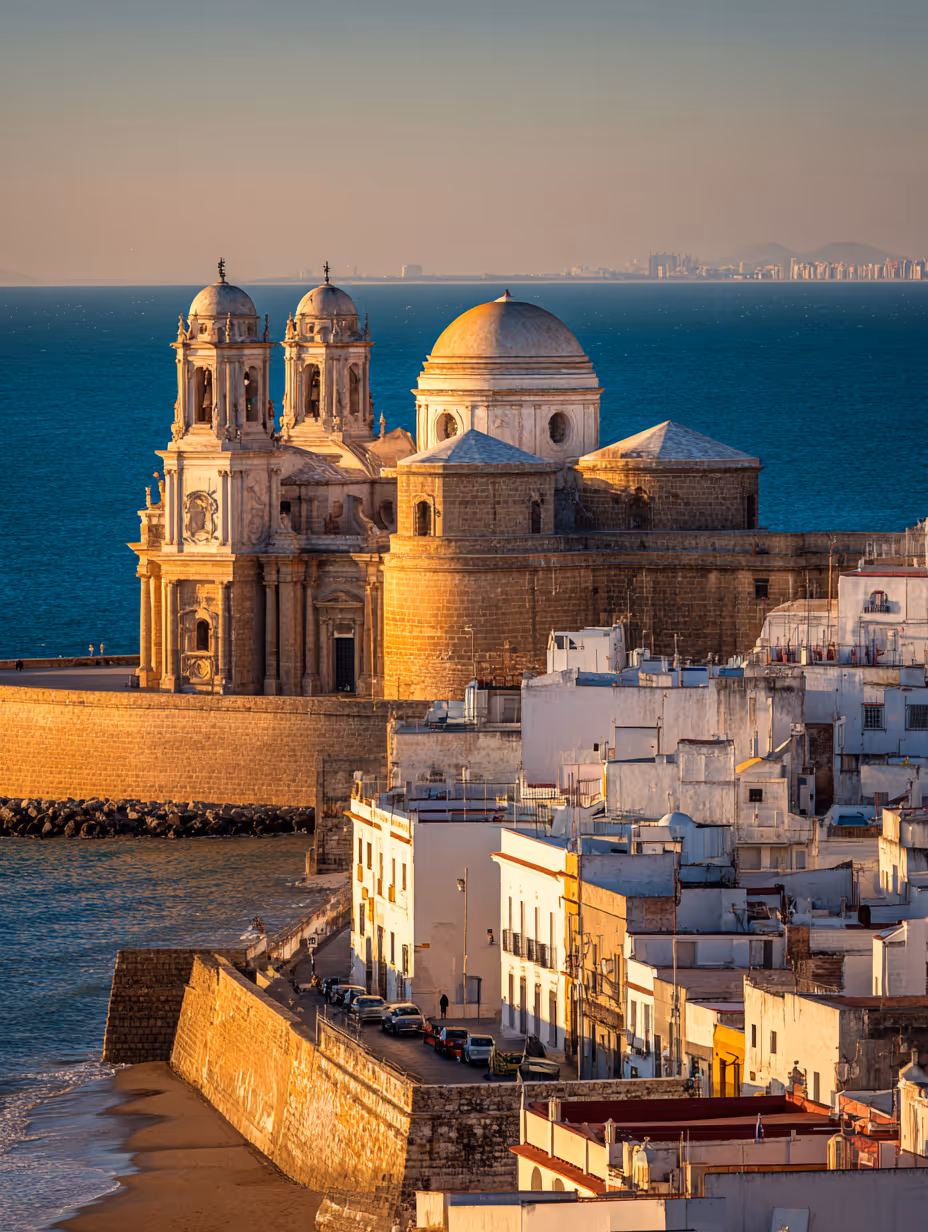 Historische kathedraal aan zee met witte gebouwen op de voorgrond bij zonsondergang.