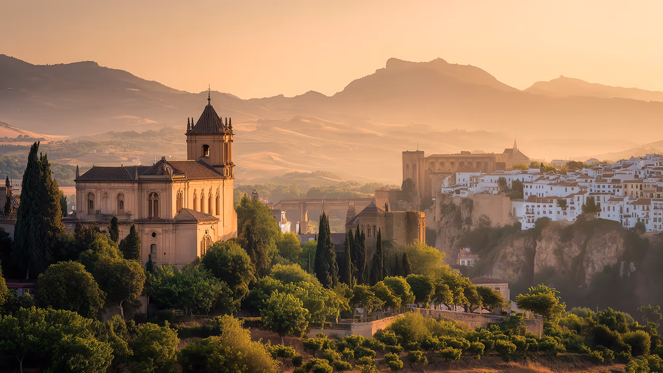 Ouderwetse stenen gebouwen en witte huizen op een heuvel met bomen en bergen in de zachte zonsondergang.