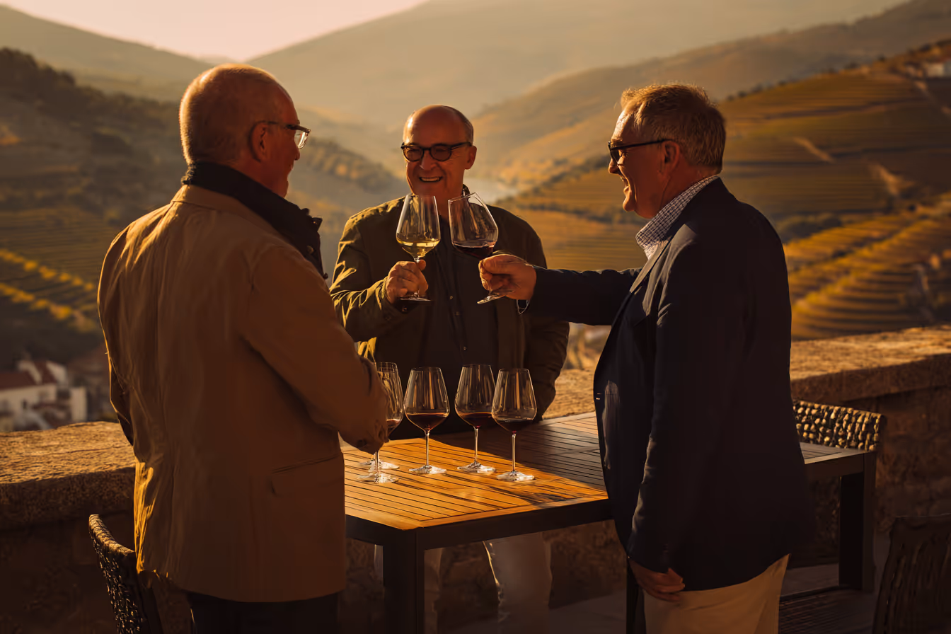 Drie oudere mannen klinken met glazen wijn bij een tafel op een terras met uitzicht op glooiende wijngaarden in de avondzon.