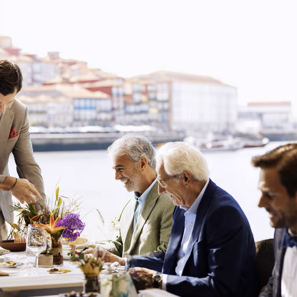 Drie oudere mannen zitten aan een gedekte tafel bij het water, terwijl een vrouw iets op de tafel legt.