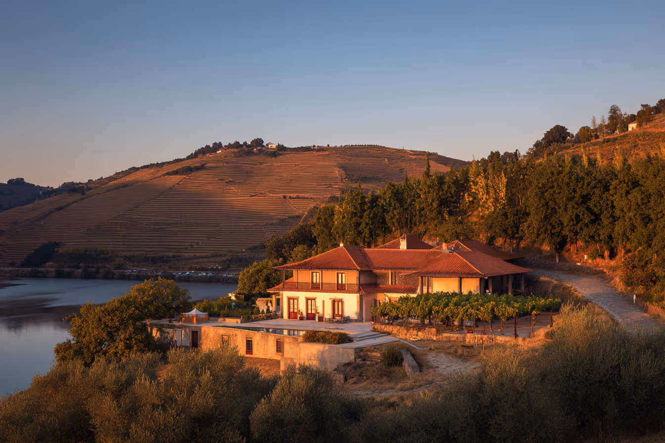 Landhuis met rood dak aan een rivier, omgeven door wijngaarden en glooiende heuvels bij zonsondergang.