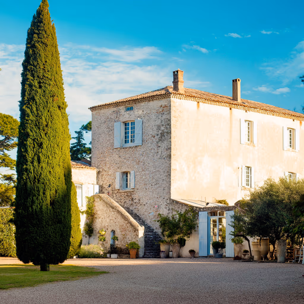 Oud stenen huis met blauwe luiken en hoge cypresboom in zonnige tuin.