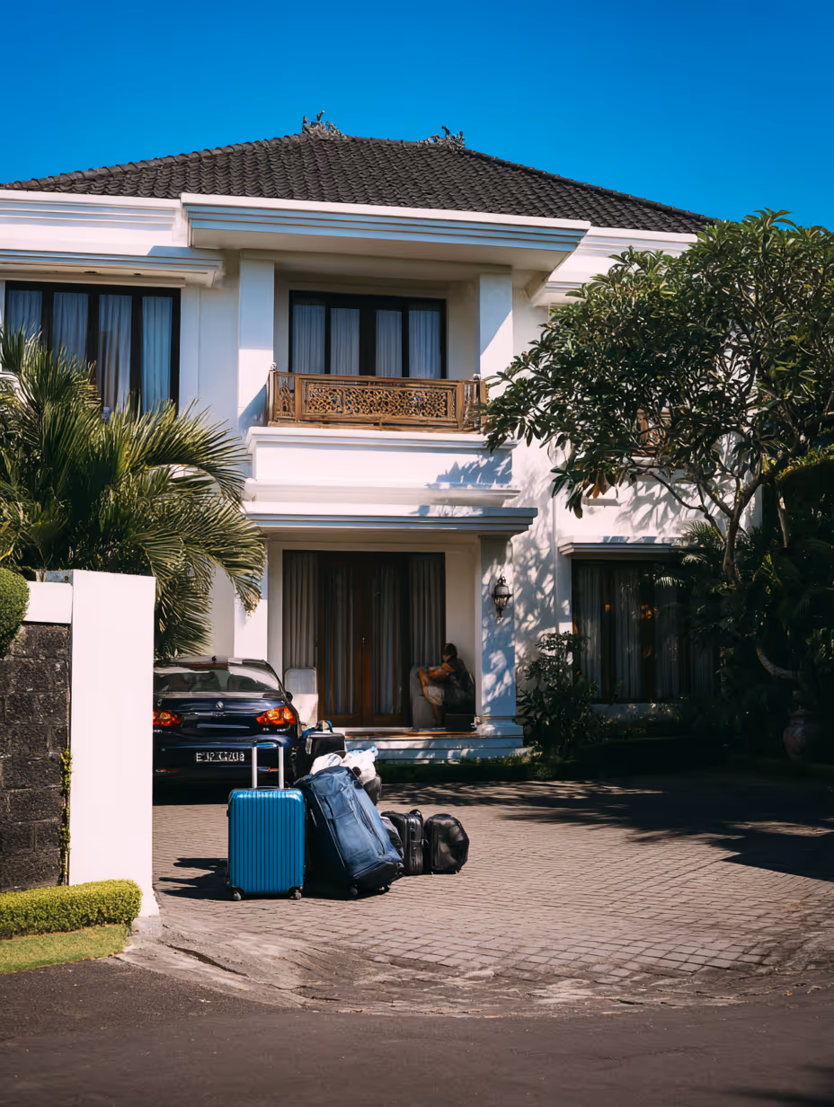 Voortuin van een wit huis met meerdere koffers op de oprit en een auto geparkeerd, met een persoon zittend bij de voordeur.
