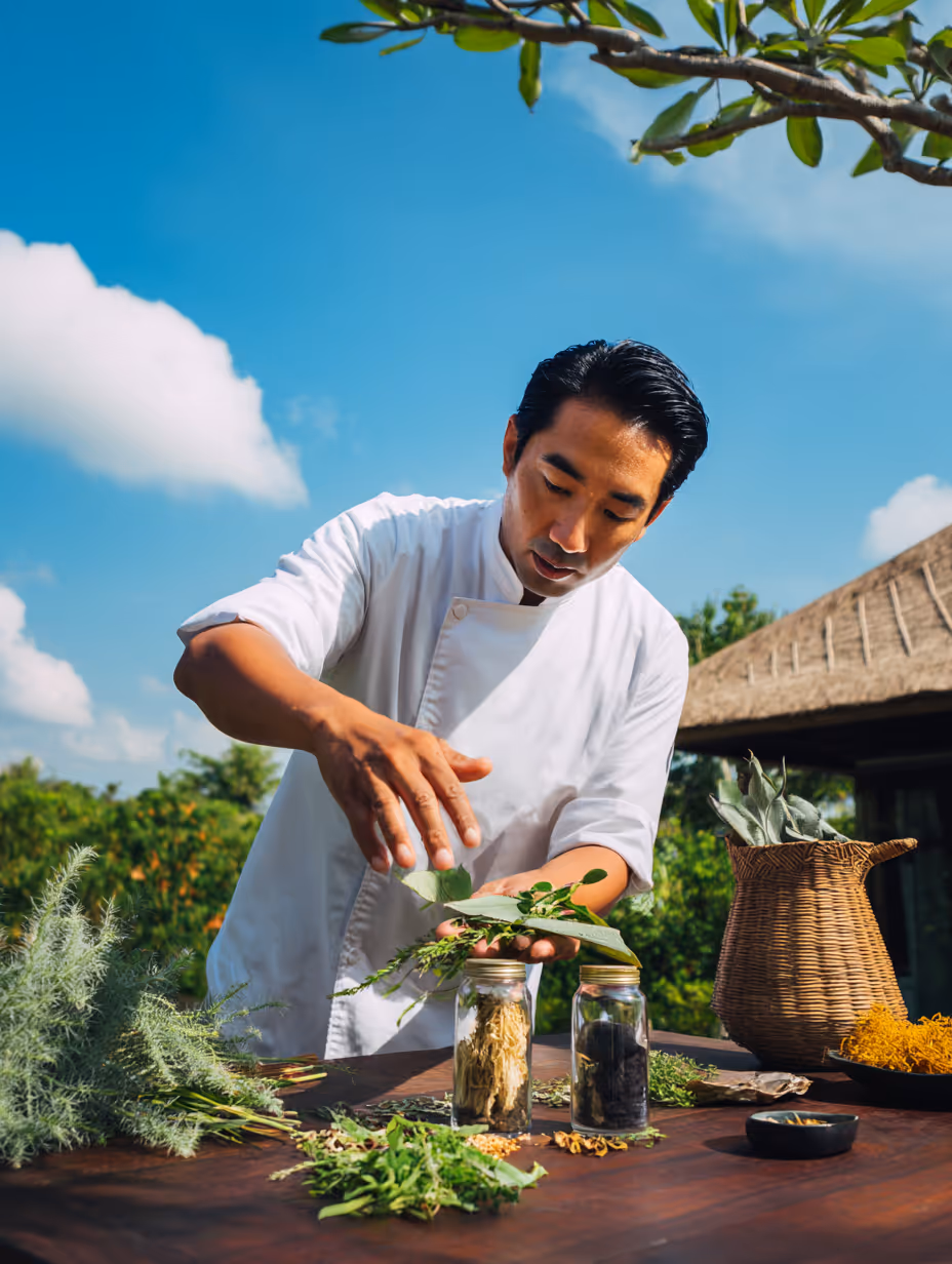 Kok in een witte jas bereidt verse kruiden op een houten tafel met glazen potten en een mand buiten onder een blauwe lucht.