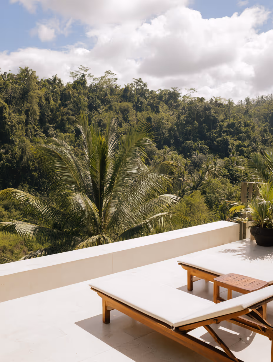 Ligbed met witte kussens op een balkon met uitzicht op weelderig groen regenwoud en palmbomen onder een partly bewolkte hemel.