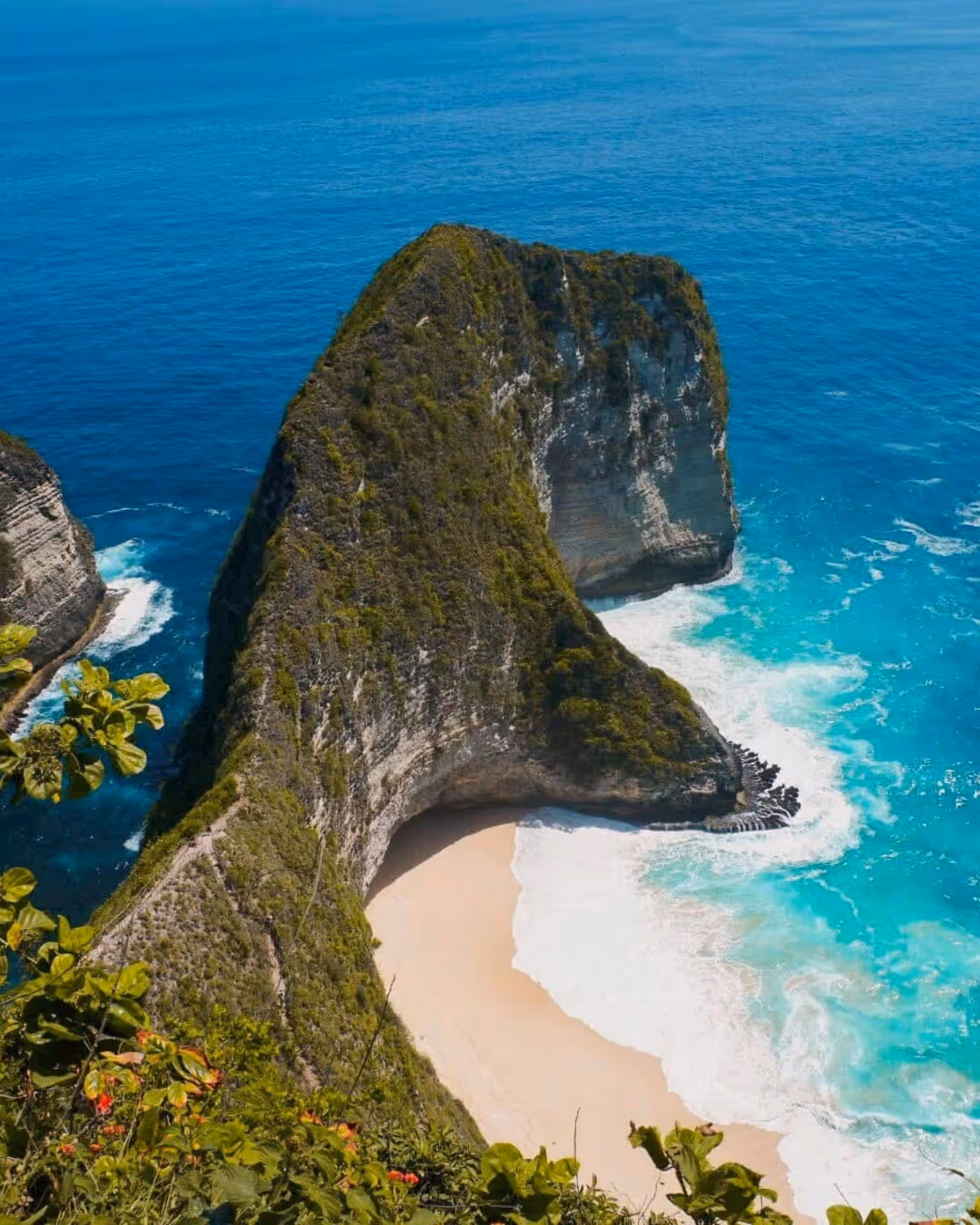 Uitzicht op een Bali  rotsachtig schiereiland met steile kliffen en een wit zandstrand, omgeven door turquoise oceaanwater en groene planten op de voorgrond.