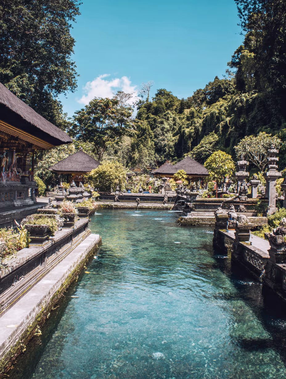 Traditioneel Balinees tempelcomplex met heldere waterpoel, weelderige groene bomen en blauwe lucht.