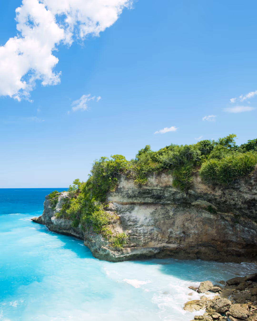 Rotsachtige Bali kustlijn met groene begroeiing en helder blauw water onder een heldere lucht met witte wolken.