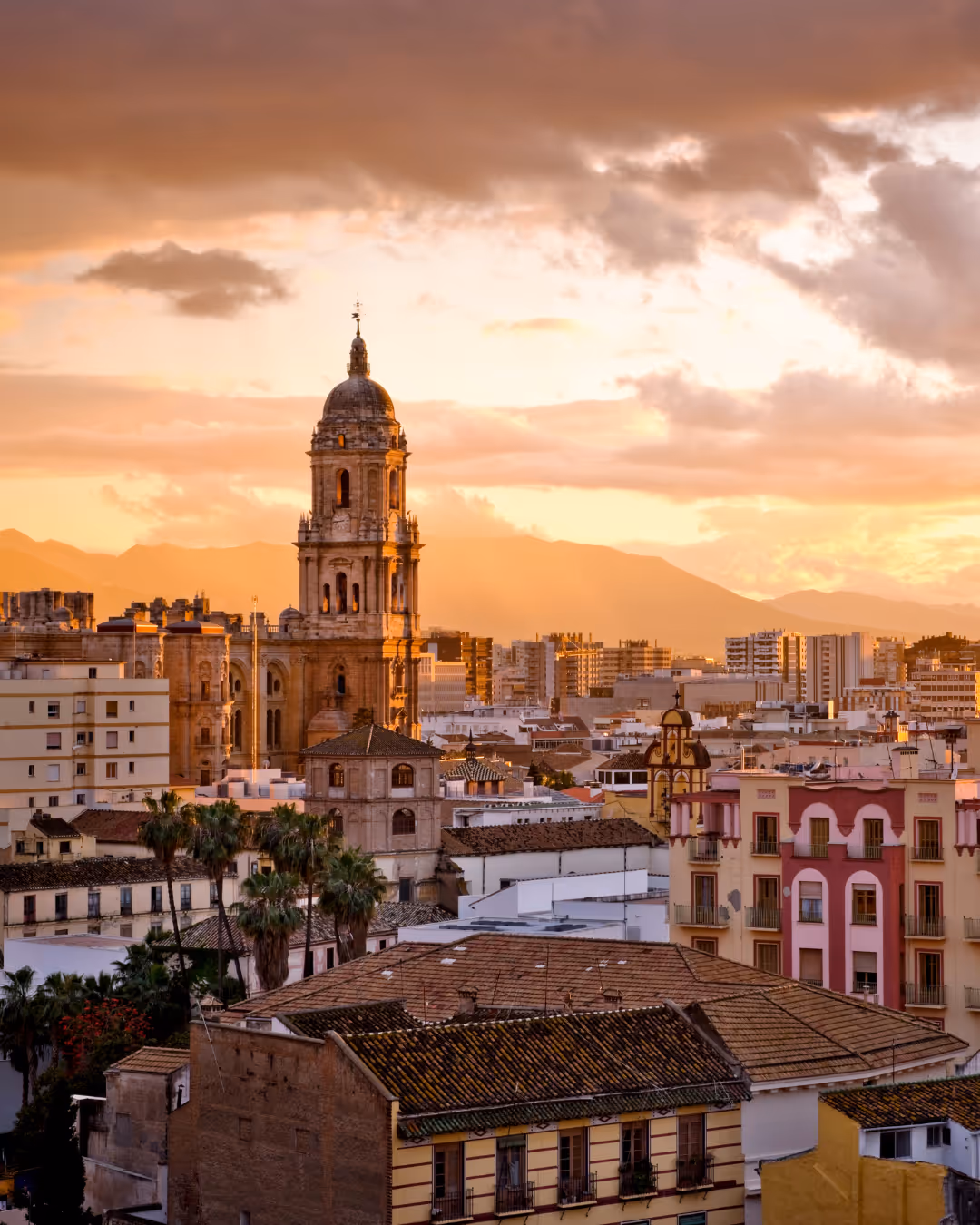 Zonsondergang boven de kathedraal van Málaga met omliggende gebouwen en bergen op de achtergrond.