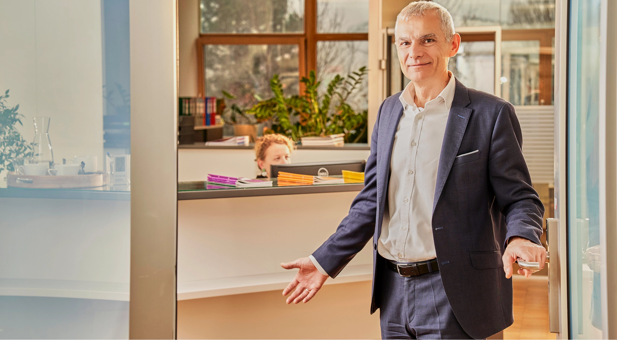 Steuerberater im Anzug öffnet die Tür und lädt in ein helles Büro mit Rezeption und einer Frau am Computer ein.