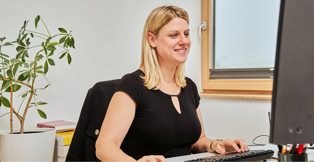 Frau mit blonden Haaren sitzt lächelnd am Schreibtisch und tippt auf einer Tastatur vor einem Monitor.