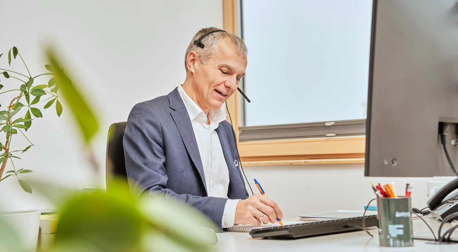 Steuerberater mit Headset sitzt am Schreibtisch, schreibt in ein Notizbuch und berät am Telefon zum Thema Finanzrecht.