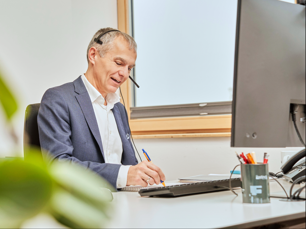 Steuerberater im Anzug mit Headset sitzt am Schreibtisch, schreibt in ein Notizbuch und blickt auf den Computerbildschirm.