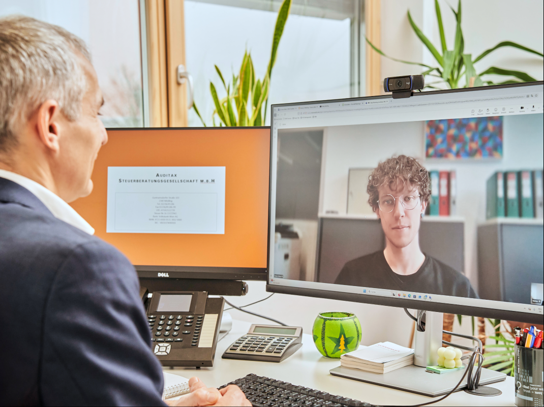 Steuerberater führt ein Videogespräch mit einem jüngeren Mann auf einem Computermonitor in einem Büro mit Pflanzen und Büroausstattung.