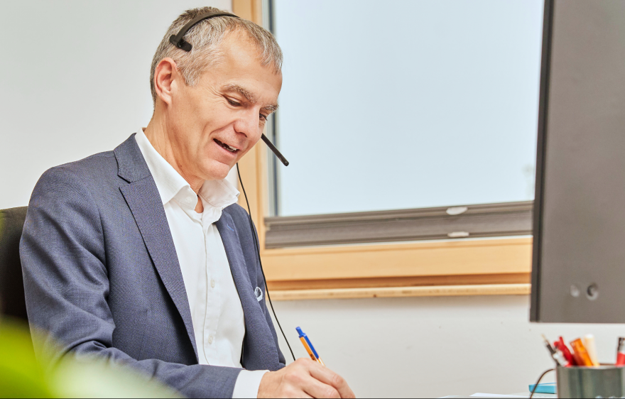 Steuerberater mit Headset sitzt am Schreibtisch und notiert etwas während eines Telefonats.