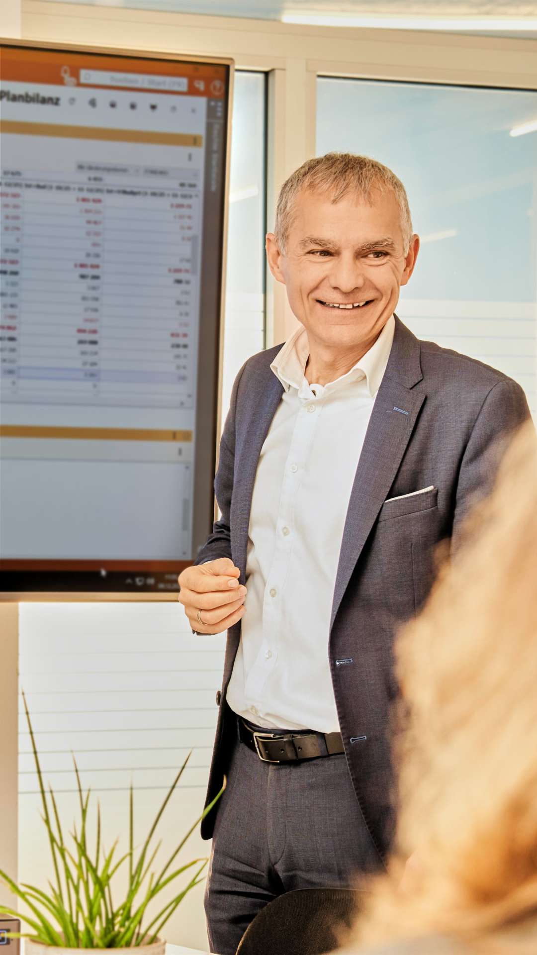 Steuerberater in grauem Anzug hält Präsentation vor Bildschirm mit Finanzdaten in auditax Büro.