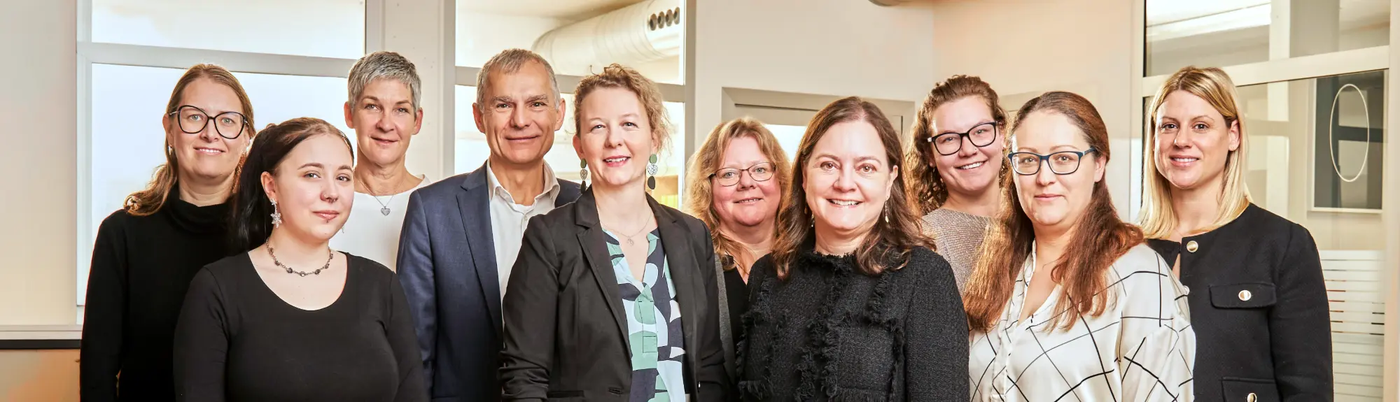 Ein Team von zehn Personen, neun Frauen und ein Mann, steht in dem hellen Büro von Auditax und lächelt in die Kamera.