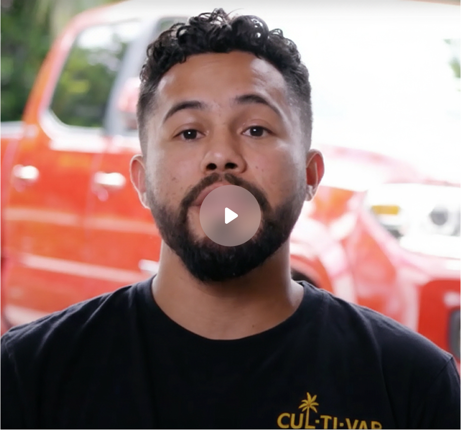Man with curly hair and beard wearing a black shirt with yellow text, standing in front of a red vehicle.