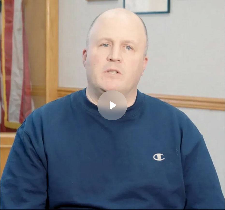 Middle-aged man in a navy blue Champion sweatshirt speaking indoors with framed pictures and an American flag in the background.