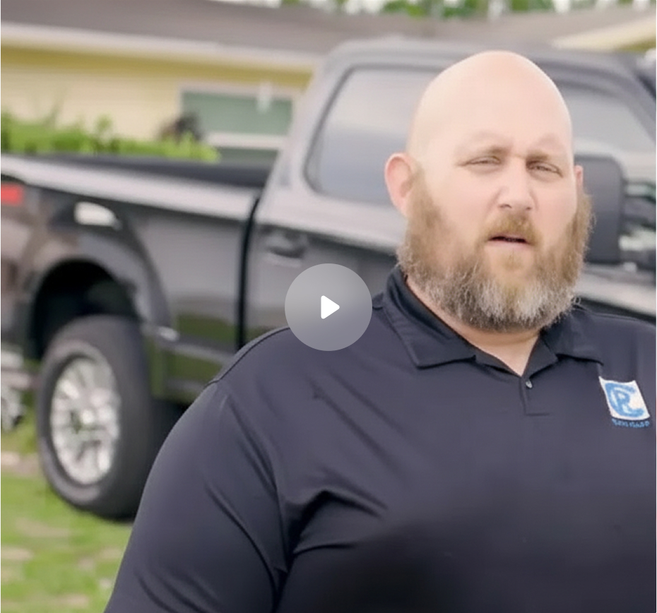 Bearded man wearing black polo shirt with logo standing in front of a black pickup truck outside a house.