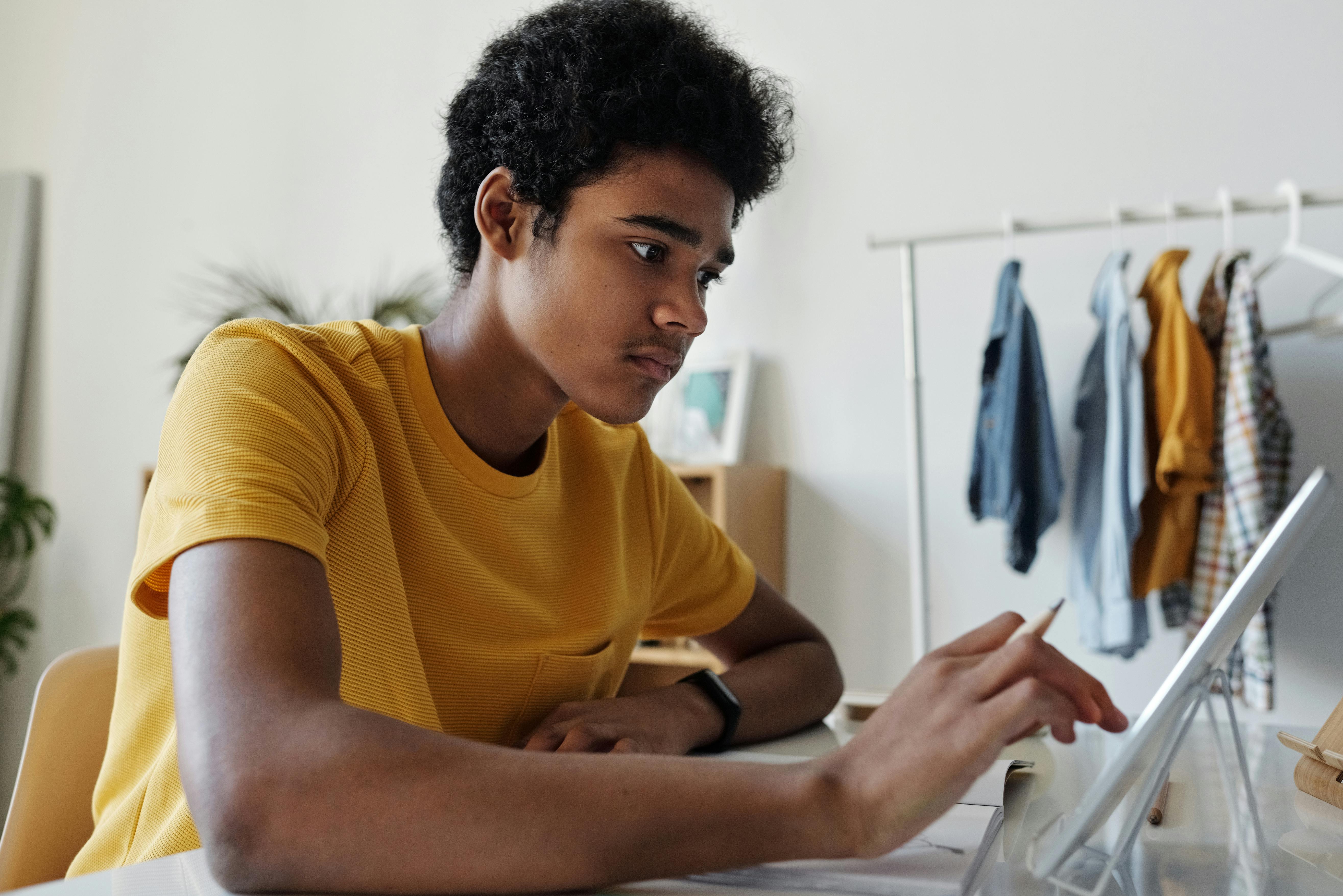 Young man in a yellow shirt focused on using a tablet with a stylus at a desk in a room with hanging clothes and plants.