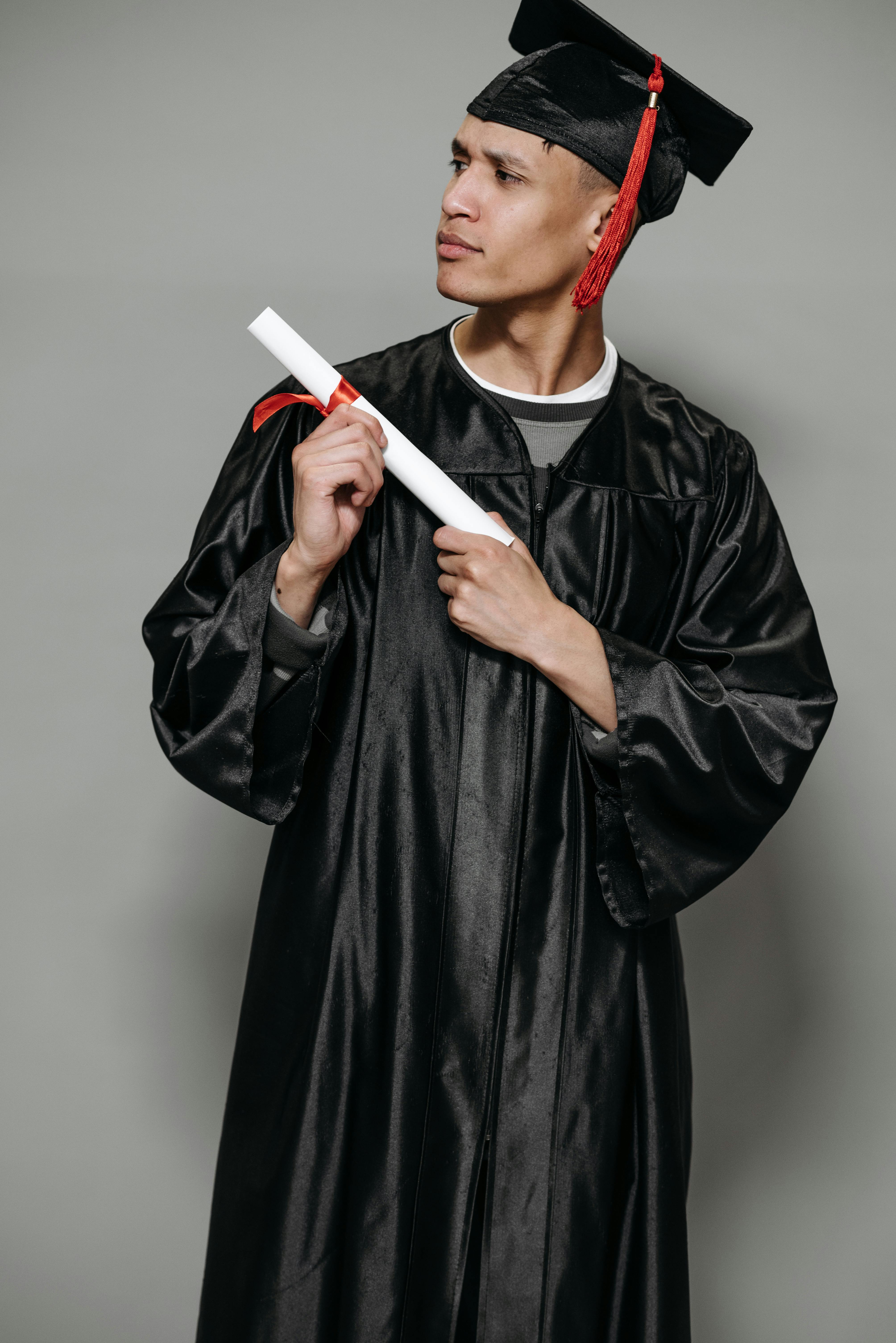 Young man wearing black graduation gown and cap holding a diploma tied with a red ribbon.